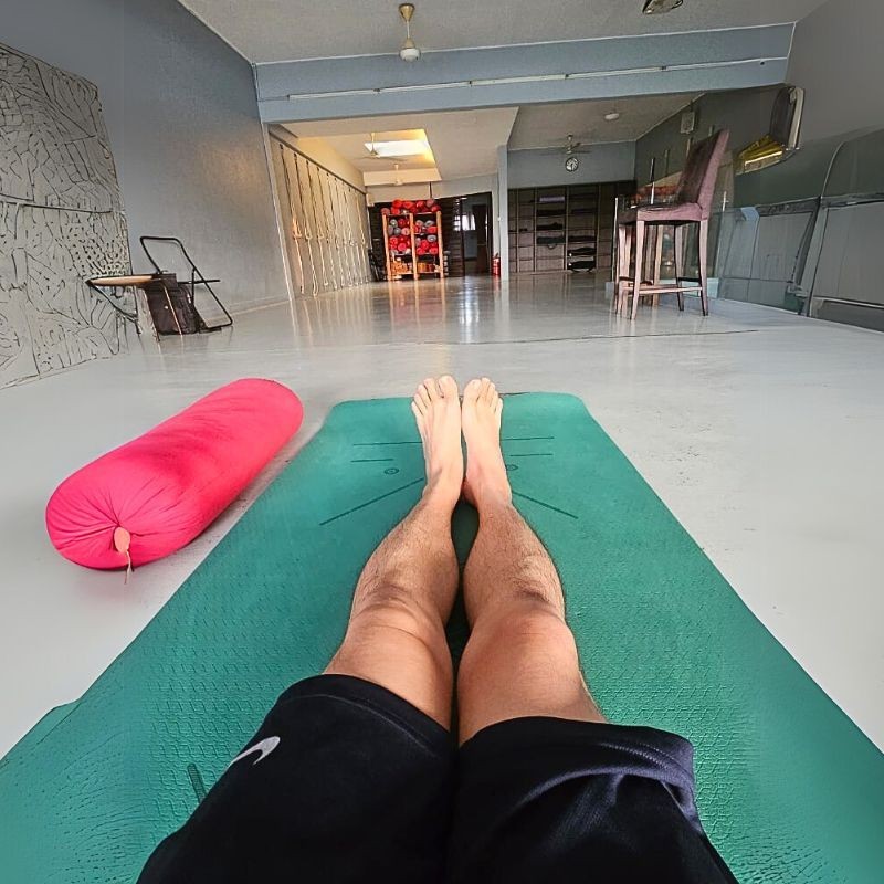 Person lying on a yoga mat in a spacious studio with a pink bolster nearby.