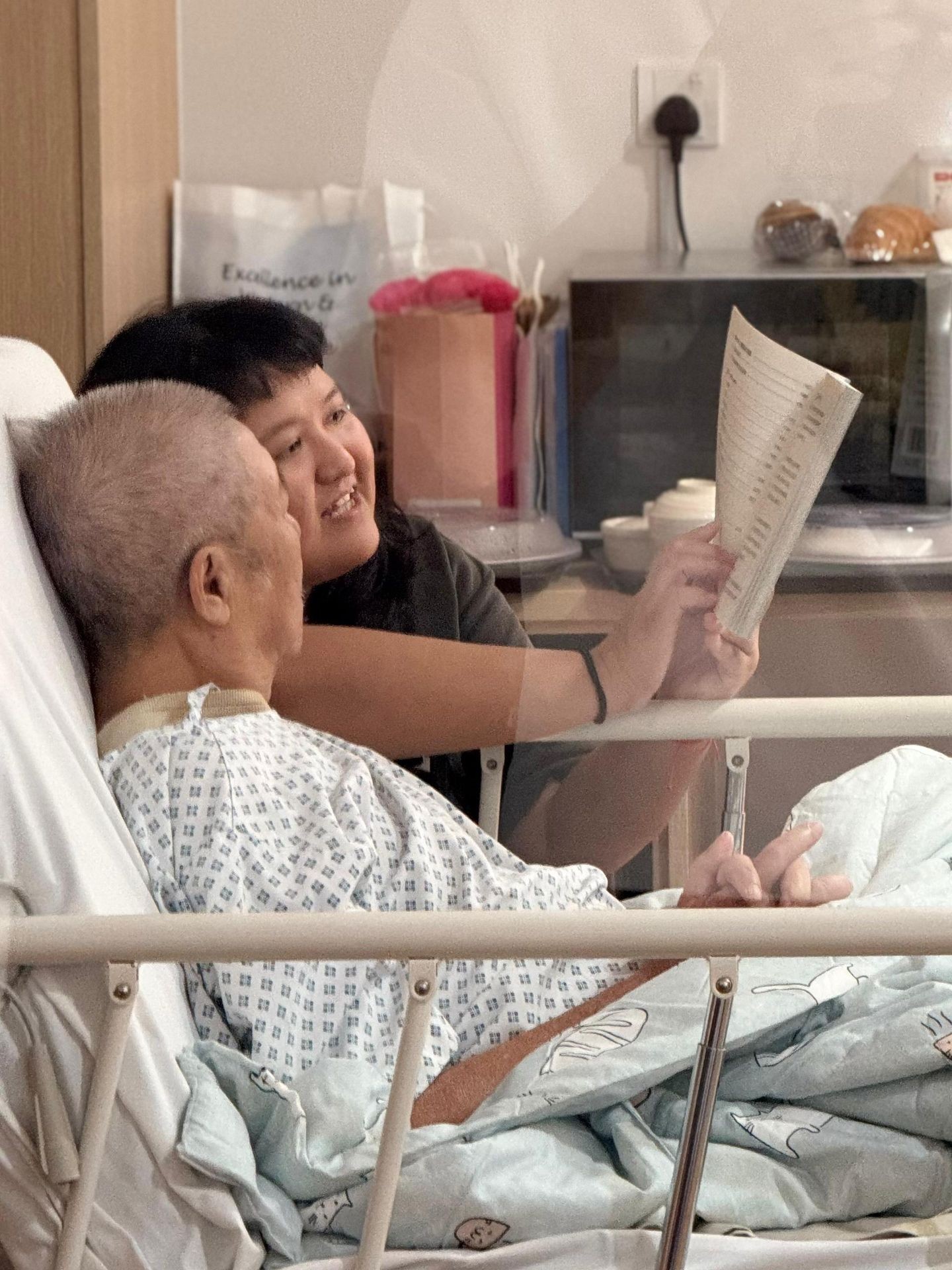 Person reading a book to a patient in a hospital bed, both appear engaged in the conversation.