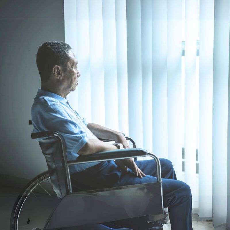 Man in a wheelchair looking out the window with vertical blinds.